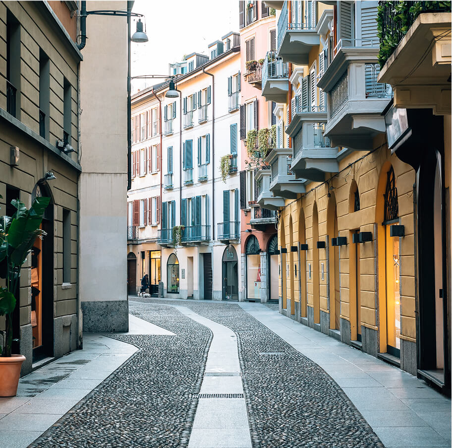 Una stretta via acciottolata di Milano fiancheggiata da edifici residenziali tradizionali con balconi e persiane colorate, e negozi al piano terra.