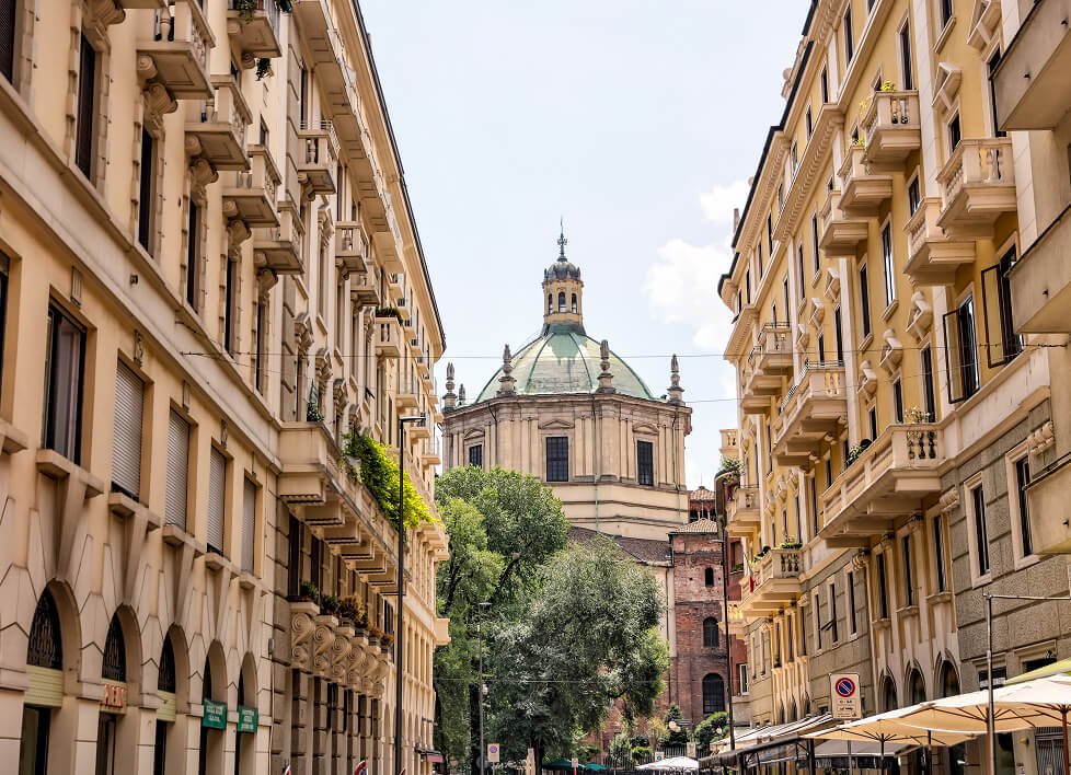 Una strada storica di Milano con palazzi eleganti e la cupola verde del Tempio Civico di San Sebastiano sullo sfondo.