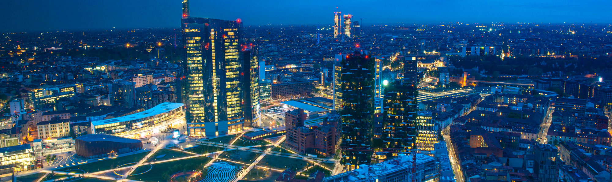 Una veduta aerea notturna dello skyline illuminato di Milano, con grattacieli moderni e un parco urbano verde illuminato.