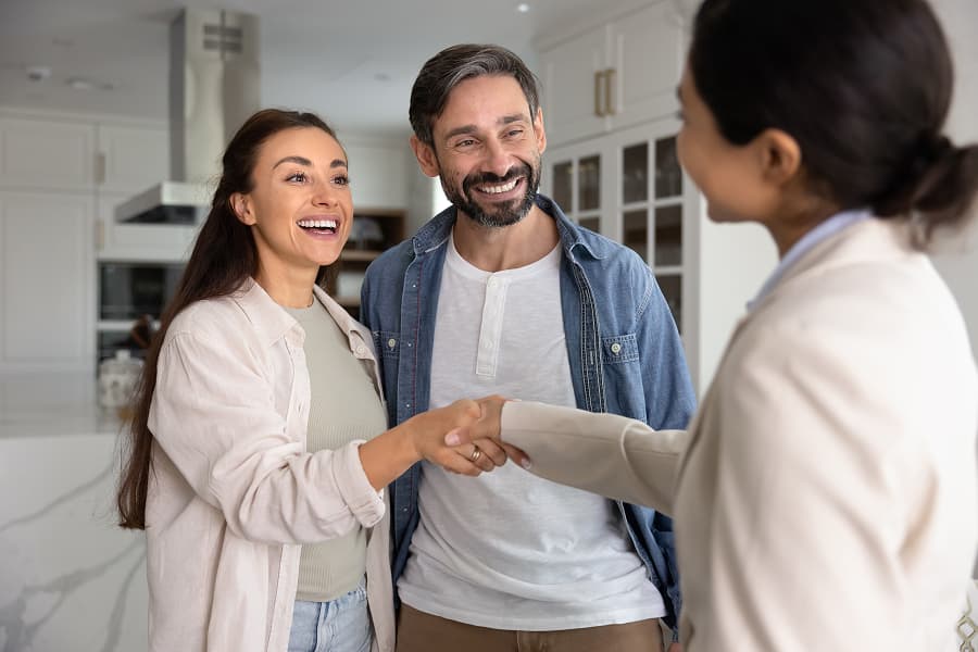 Coppia sorridente stringe la mano a una professionista in un ambiente domestico, aria di benvenuto e cordialità.