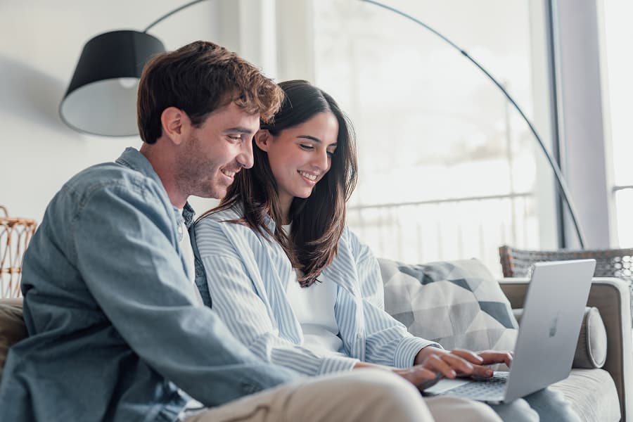 A happy couple sitting on the sofa at home using a laptop, with satisfied smiles, in a bright, modern setting with minimalist furnishings and large windows
