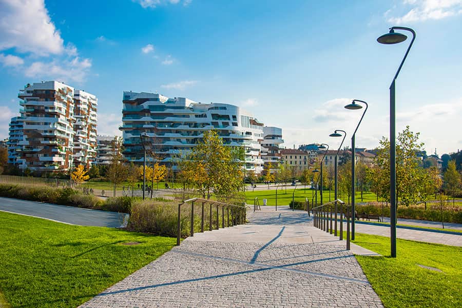 Immagine di un parco moderno con percorso pedonale, illuminazione pubblica e edifici innovativi sullo sfondo, in una giornata soleggiata primaverile