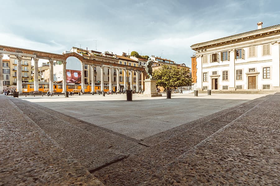 Immagine delle Colonne di San Lorenzo con archi storici e edifici eleganti, caratterizzata da ampio spazio aperto, alberi e una scena urbana tranquilla, ideale per passeggiate e socializzazione.