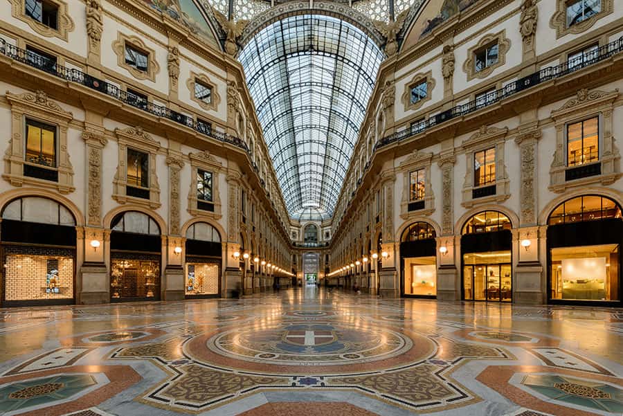 Interno della galleria Vittorio Emanuele con soffitto in vetro, pavimento decorato, luci calde e negozi di lusso ai lati, creando un'atmosfera sofisticata e spaziosa