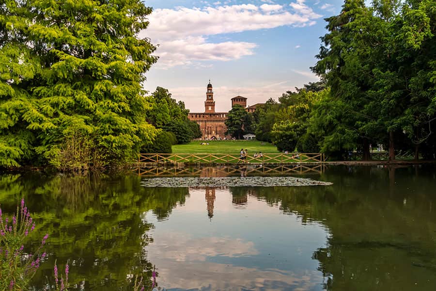 Immagine del Parco Castello con un lago che riflette il cielo sereno e il Castello, circondato da alberi verdi e un sentiero di legno, sotto un cielo leggermente nuvoloso.