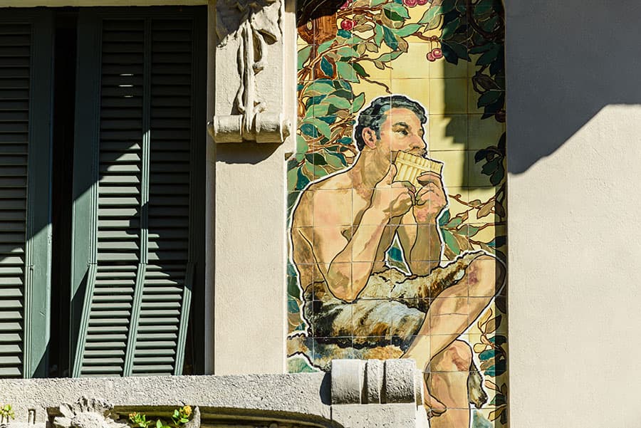 Murale raffigurante un uomo senza maglietta seduto, che sorseggia una bevanda, sfondo di piante e foglie di canto, affacciata su una parete esterna di edificio, con dettagli in ombra e luce naturale.
