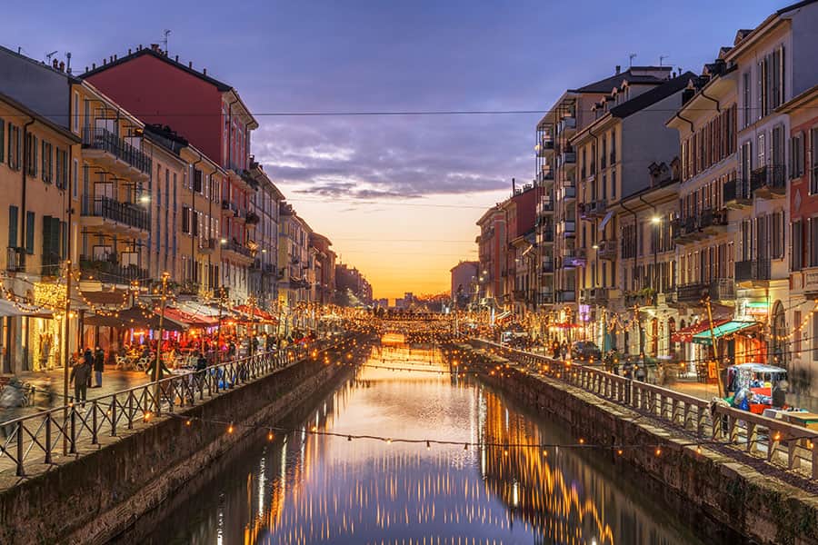 Veduta vivace dei Navigli con edifici colorati e negozi illuminati, al tramonto, atmosfera festosa e affollata, riflessi dell'illuminazione sull'acqua