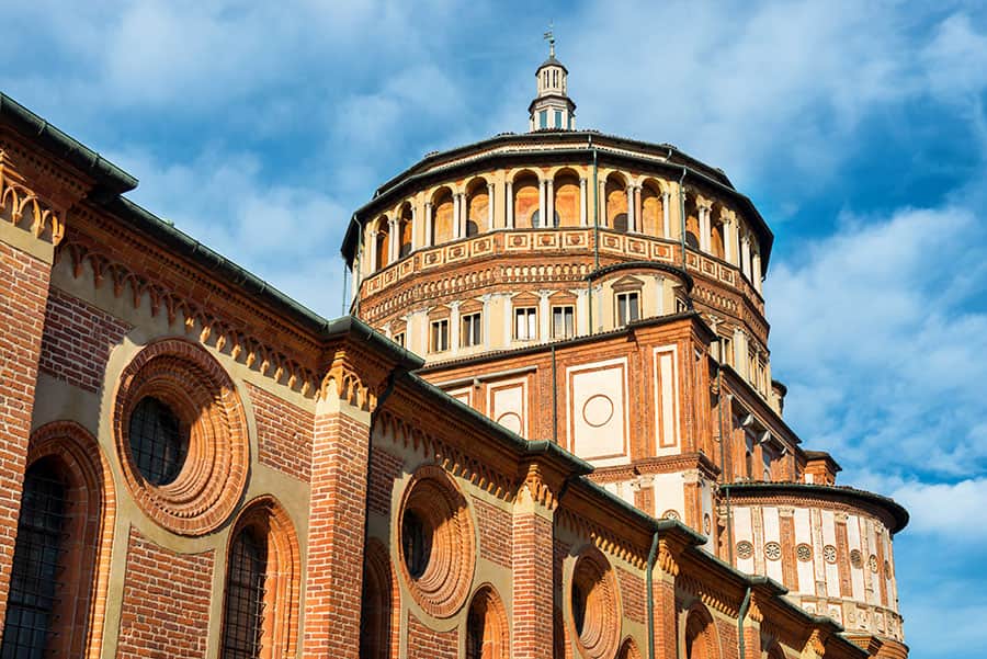 Immagine di una chiesa storica con dettagli in mattoni rossi, grandi finestre ad arco e una cupola imponente sotto un cielo sereno, rappresentativa dell'architettura religiosa italiana.